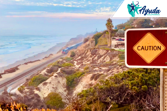 A “Caution” sign stands before coastal cliffs in California, with a train passing in the background under cloudy skies, symbolizing the increased landslide risks after recent Southern California rains. The image highlights Ayuda California’s commitment to prevention and support for affected communities.