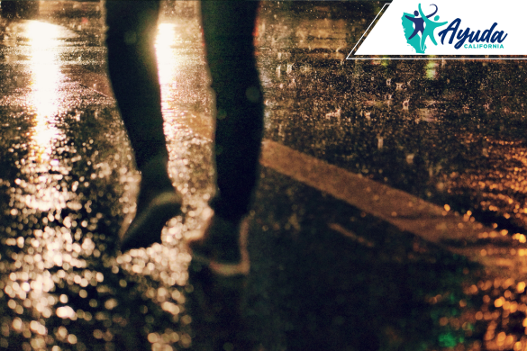 A person walks on a wet street illuminated by city lights during a rainy night, symbolizing the vulnerability of pedestrians in hazardous weather conditions. The image reflects the fatal pedestrian accident in Fresno during the rain, as reported by Ayuda California.