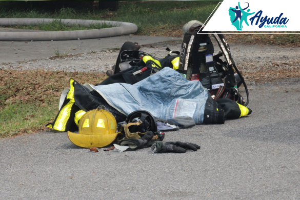 Firefighter gear including a helmet, gloves, and jacket lies on the ground next to a hose, symbolizing the seriousness of a hit-and-run incident while battling a truck fire in Bakersfield. The image highlights the vulnerability of emergency responders on California highways, as reported by Ayuda California.